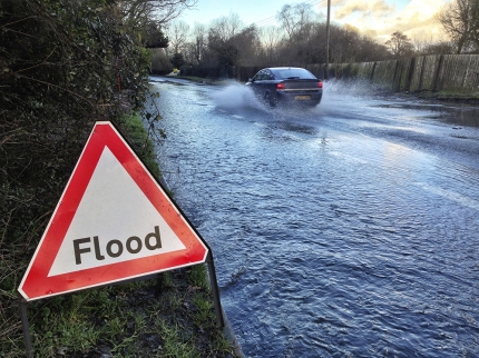 A flood warning sign and a car passing through a flooded road behind