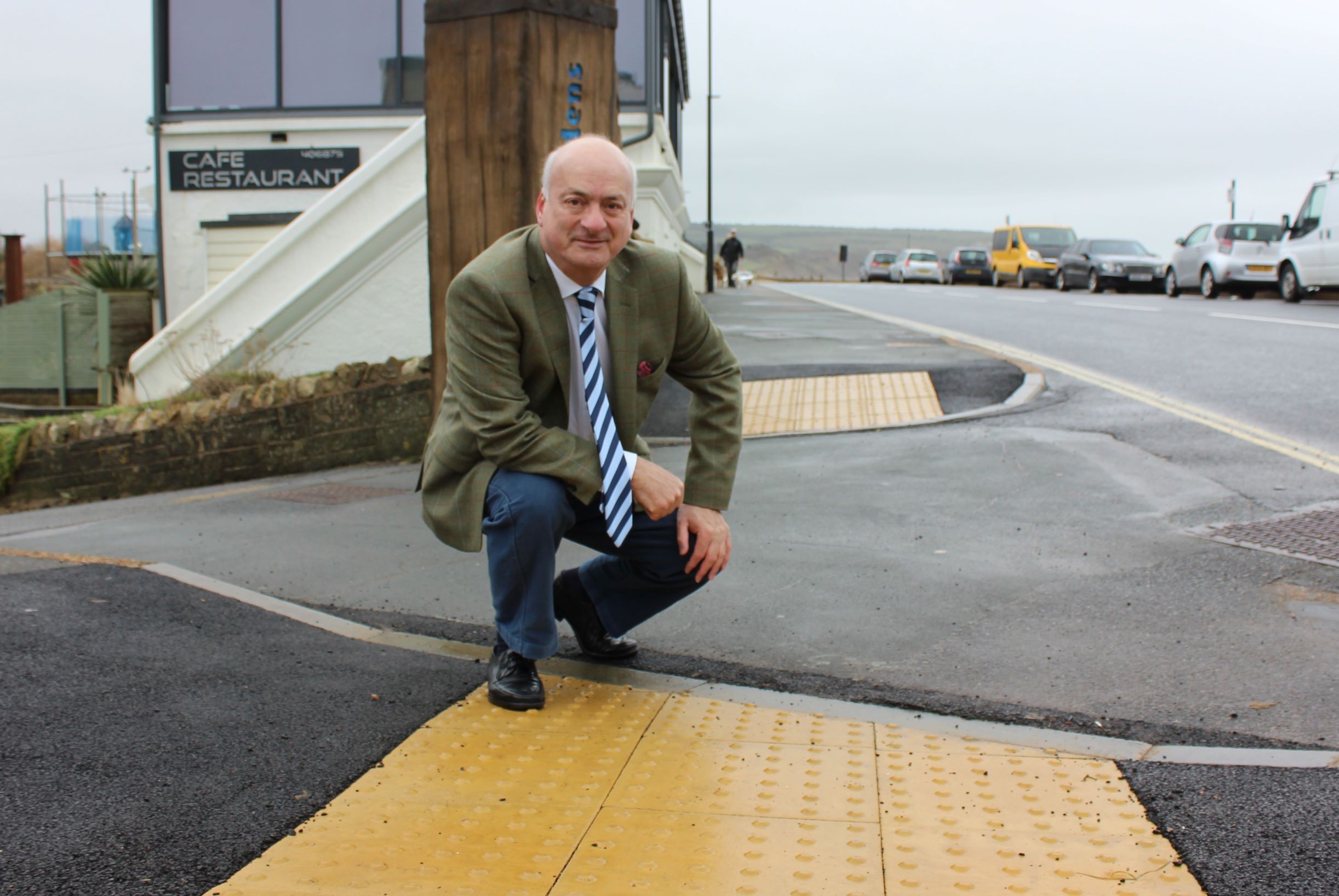 Photo shows a person (councillor Ian Ward) crouched down beside a tactile dropped kerb near the edge of the road