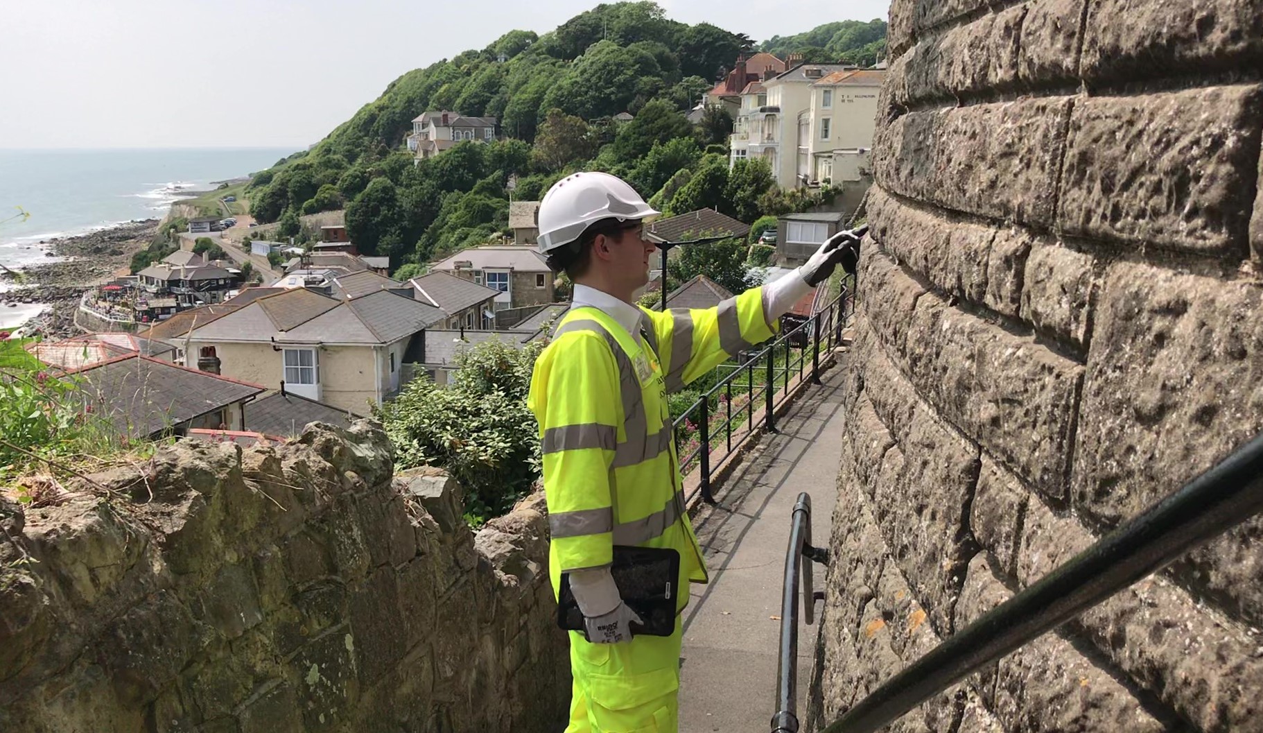 Photo showing a man in protective uniform (PPE) inspecting a wall with the town of Ventnor and seaside in the background