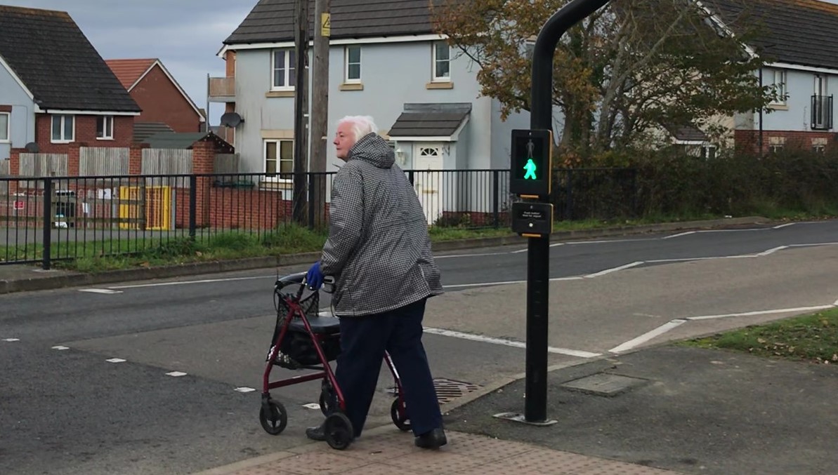 Older lady with walking aid crossing from tactile pavement at a pedestrian crossing housing estate in the background