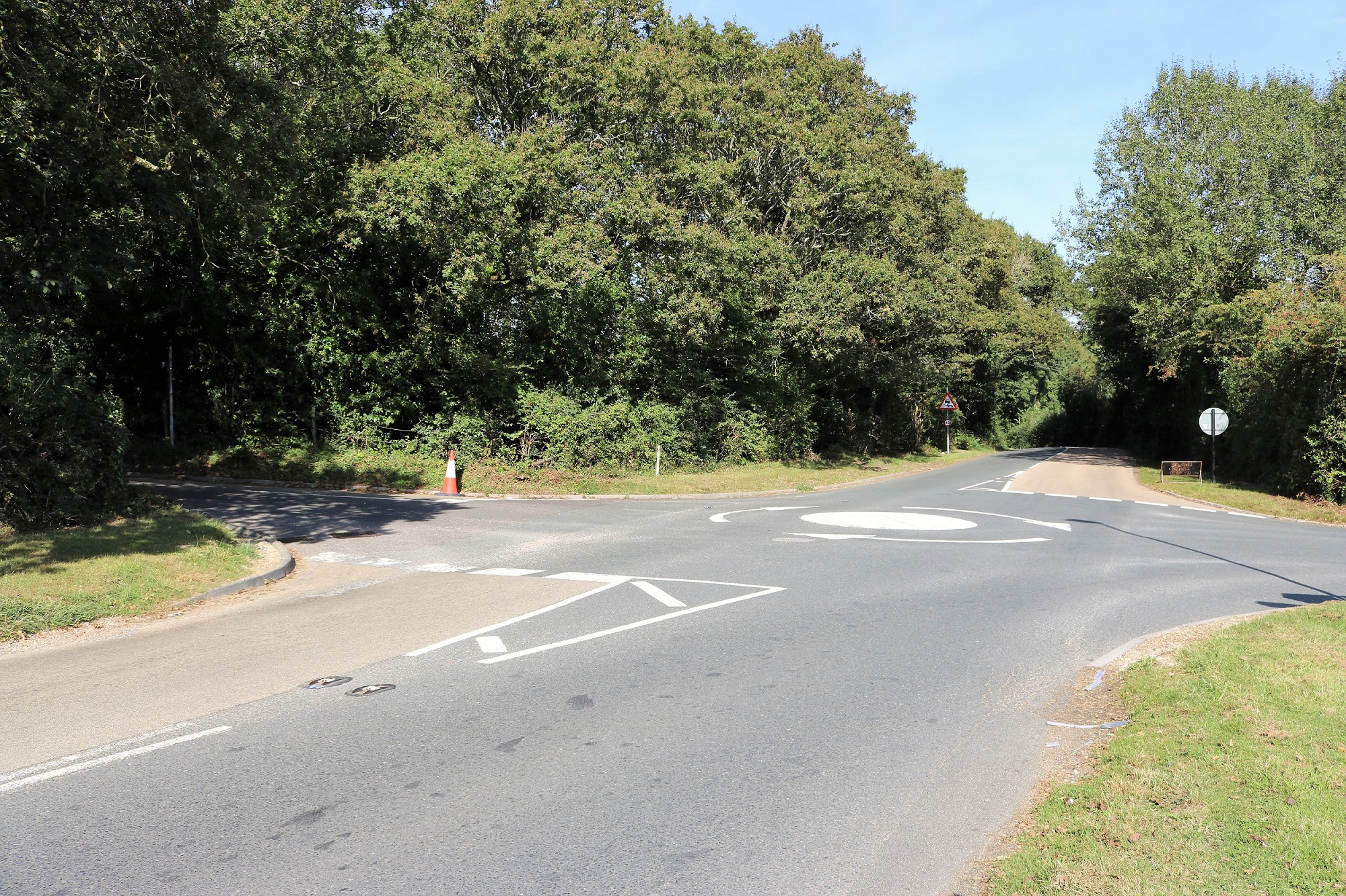 Four way road junction with mini roundabout, trees and hedging bordering the roadside