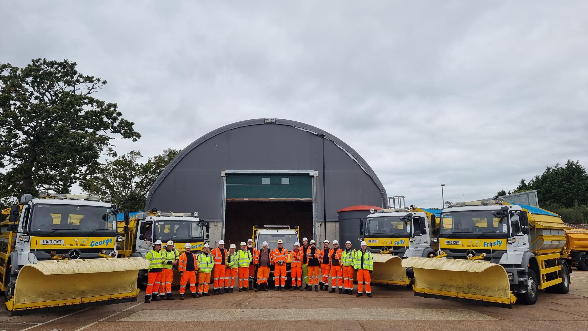 several gritting lorries lined up in a semi circle facing with a large group of men in hard hats and brightly coloured protective work gear. Large storage barn in the background and trees in the distance either side.