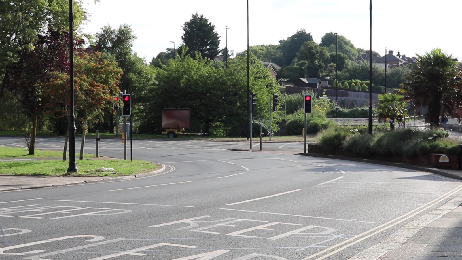 three lane road with traffic lights on red at the junction with Coppins Bridge roundabout, greenery and trees in the background and some houses