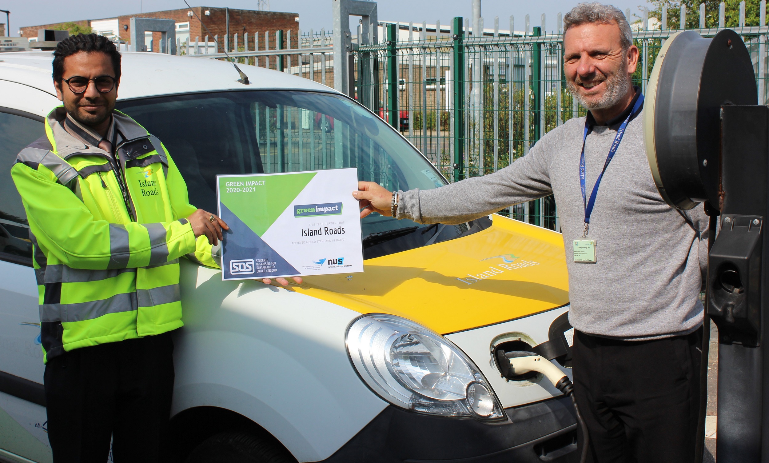 Nick Bhatnagar and Stephen Ambrose standing alongside an Island Roads electric vehicle holding the Green Impact gold award certificate