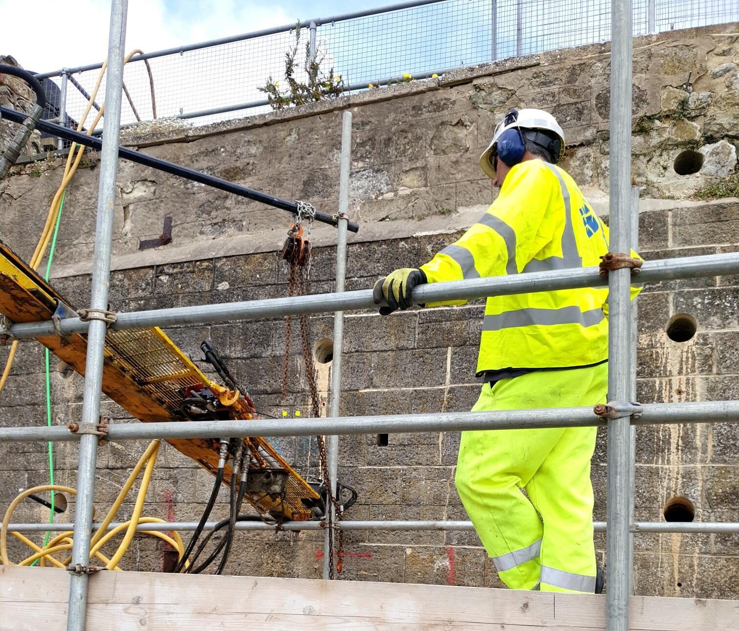 man in yellow hi visibility workwear standing on scaffolding adjacent to wall, other equipment visible in the background