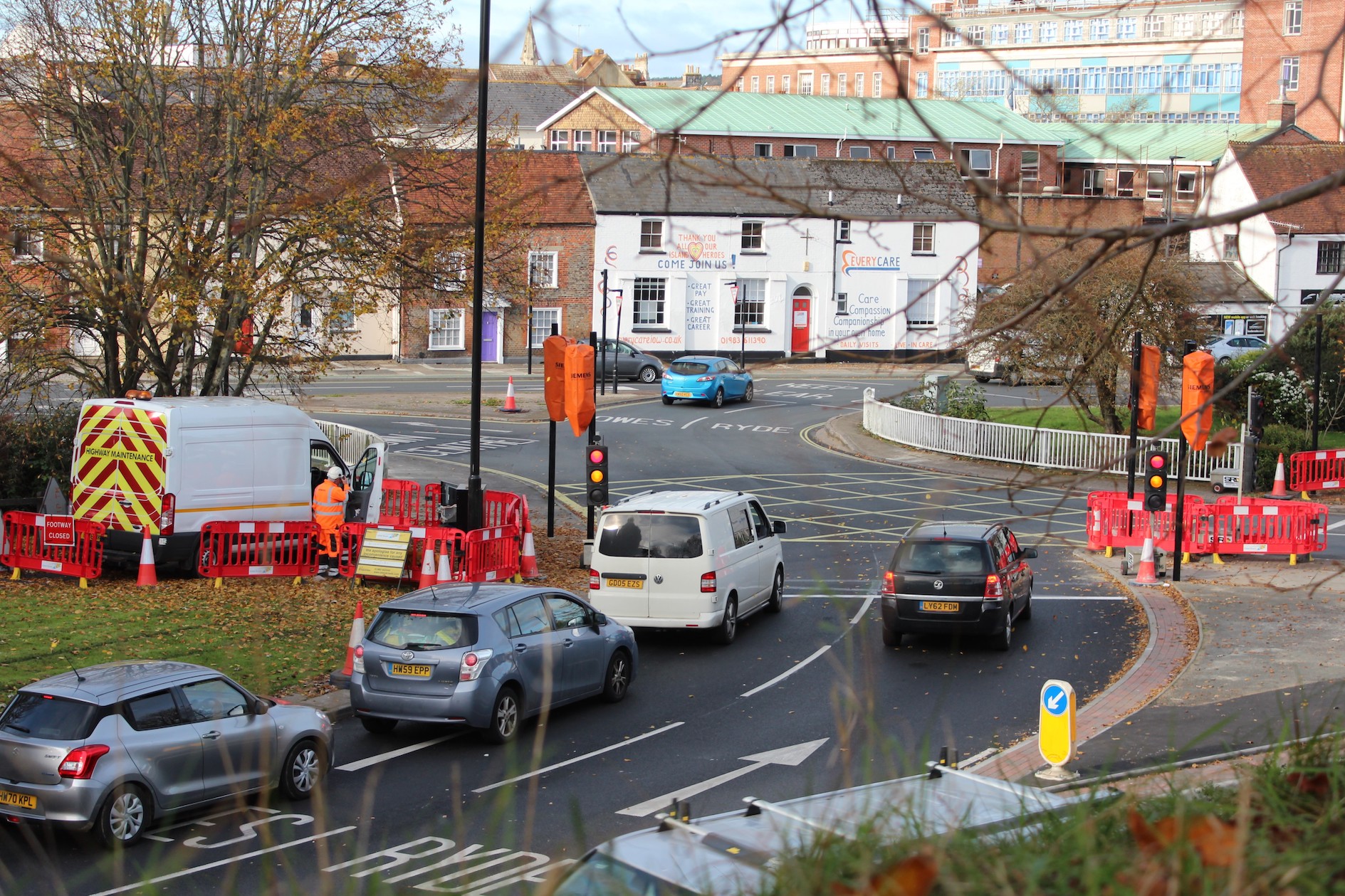 cars queuing at roundabout junction with workmen, red and white traffic barriers and signs indicating work taking place on lights. Traffic lights covered over with hoods and temporary lights in place. Buildings and trees in the background.