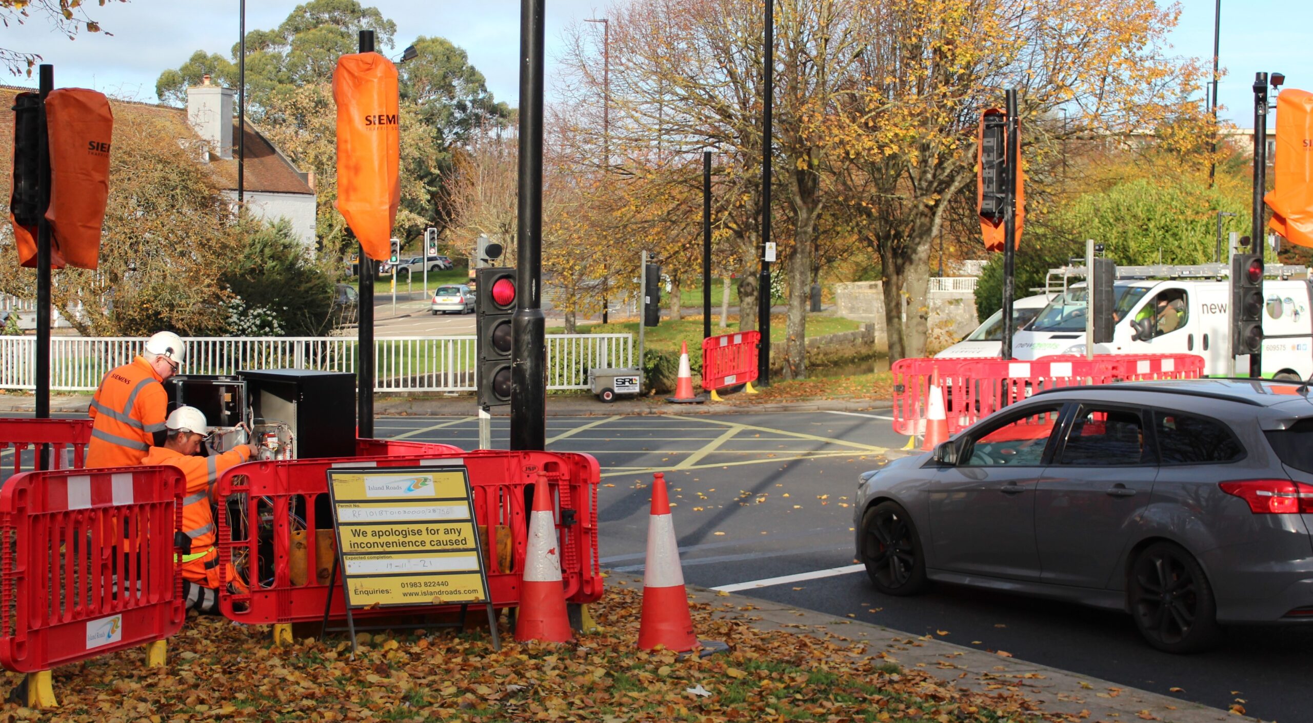 car stationary at roundabout traffic lights, other vehicles stationary on approach to the right, some traffic lights covered over with orange hoods, temporary traffic lights visible, two workman working on traffic signal box, leaves, traffic cones, signs and barriers in foreground, trees and houses in background