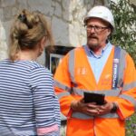 Man in hard hat and protective clothing holding a tablet and talking to a lady outside her house.