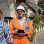 Island Roads staff member in hard hat and protective clothing carrying an iPad and talking to a member of the public in the street next to some cottages