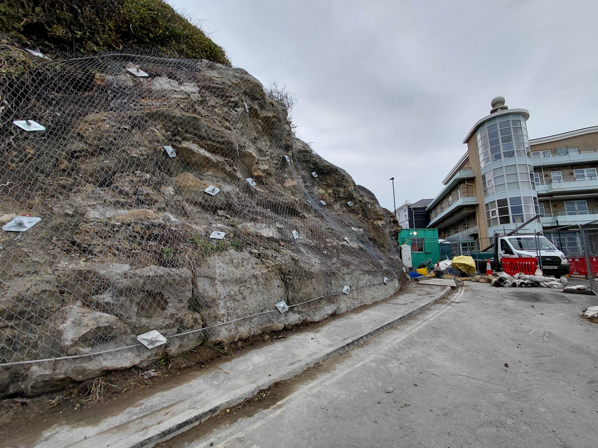 unsurfaced road with rock face to the left covered with netting. A work van can is parked at the end of the road