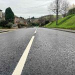 picture of a newly resurfaced road with white line down centre. Sloping grass verge on right hand side and houses on the left.