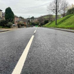 picture of a newly resurfaced road with white line down centre. Sloping grass verge on right hand side and houses on the left.