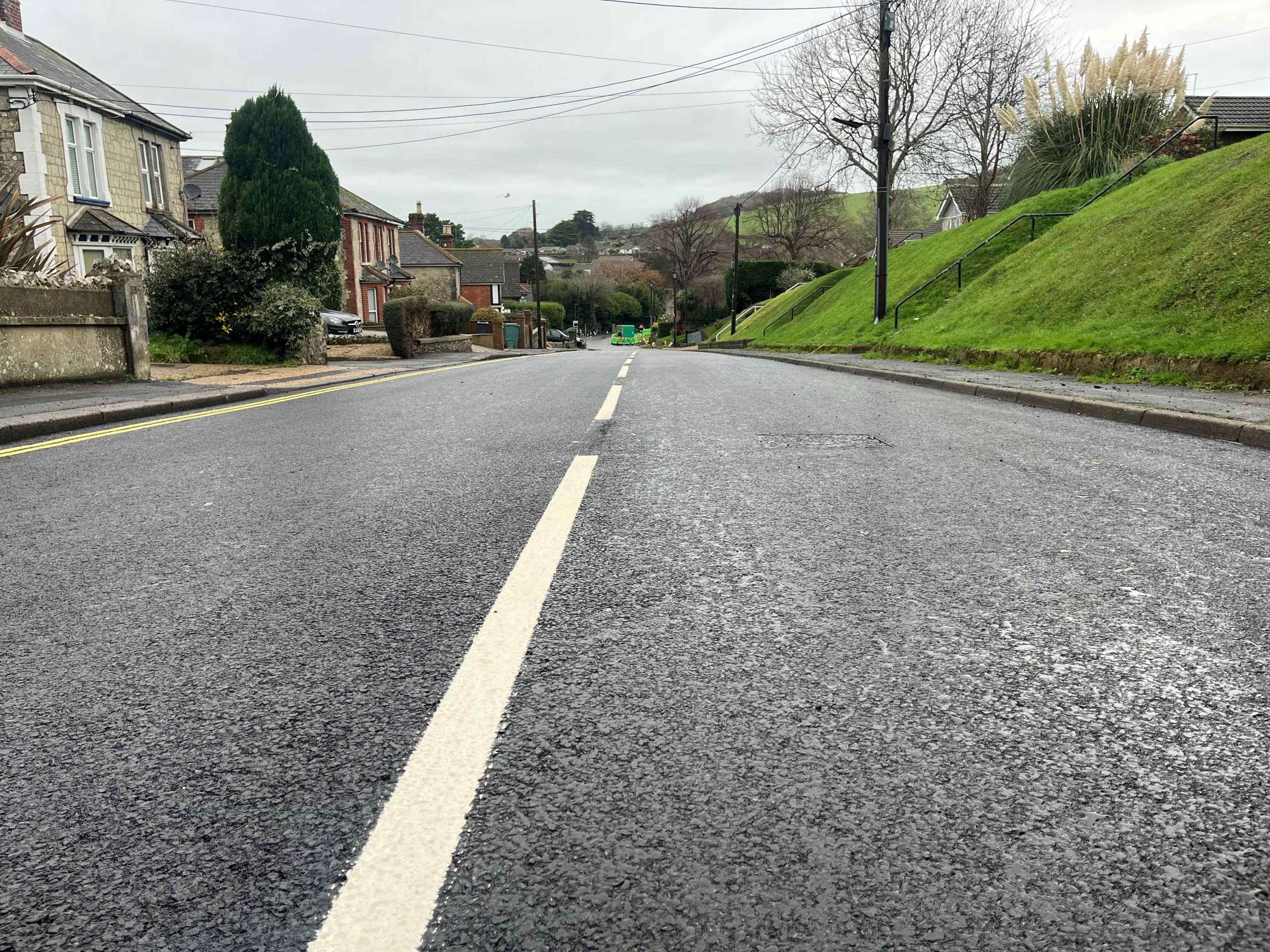 picture of a newly resurfaced road with white line down centre. Sloping grass verge on right hand side and houses on the left.