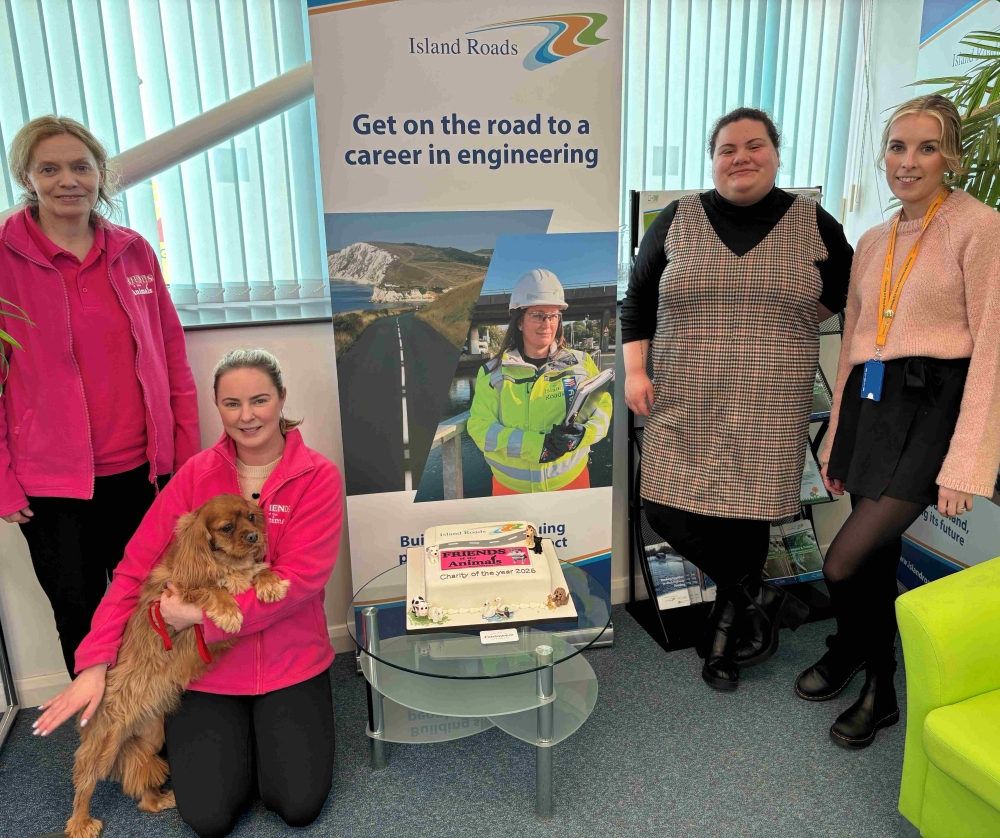 two members of Island Roads staff and two people from friends of the animals holding a dog in front of an Island Roads banner which says Get on the road to a career in engineering. There is a cake on a table which has Island Roads and friends of the animals written on it.