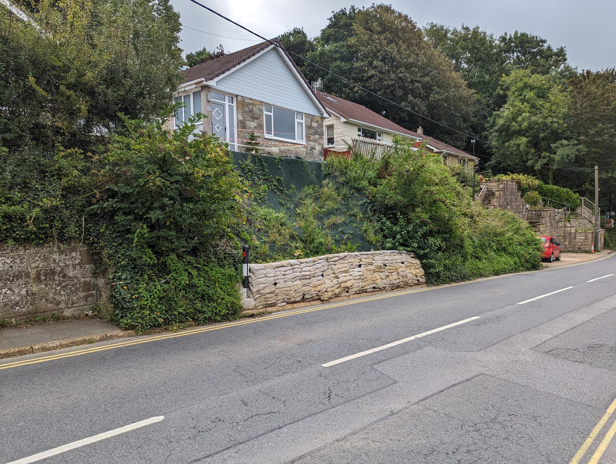 section of road, bank/verge is shored up with a 'wall' of sandbags