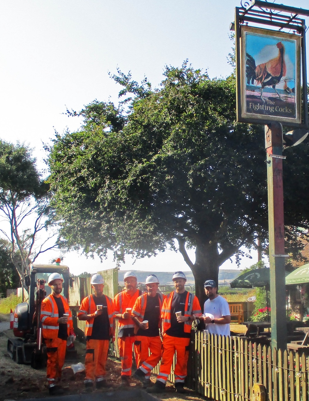 Roadworkers in hard hats and orange protective clothing standing outside a pub with the landlord and holding soft drinks.